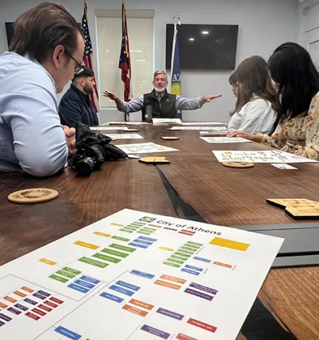 Five students, seated listen to Athens, Ohio, Mayor Steve Patterson, who is gesturing at the head of the table with a city organizational chart in the foreground.