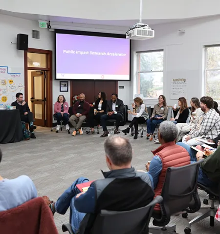 15 faculty members sit in a half circle in a conference room with a slide that says “Public Impact Research Accelerator” showing in the background.