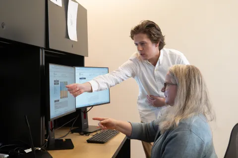 One person seated and one standing in an office point to two computer screens showing data graphs